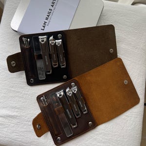 Op de afbeelding: Twee lederen manicure-sets in donkerbruin en bruin, elk voorzien van een metalen drukknoopsluiting en zilverkleurige roestvrijstalen gereedschappen zoals nagelknippers, een nagelvijl en een pincet. De gereedschappen zijn voorzien van het woord Manicure.