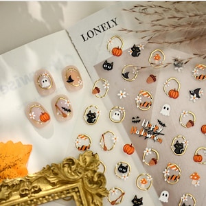 May include: A sheet of Halloween-themed nail stickers featuring small gold-rimmed icons of pumpkins, ghosts, black cats, acorns, and mushrooms, alongside the text Happy Halloween. Four individual nail tips display these designs.