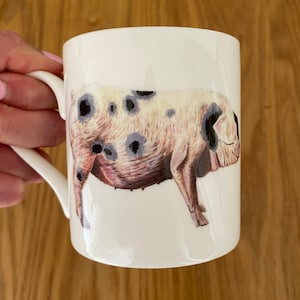 Può includere: Tazza in ceramica bianca con manico nero che presenta un'illustrazione disegnata a mano di un maiale macchiato. Il maiale è rosa con macchie nere e guarda a destra.