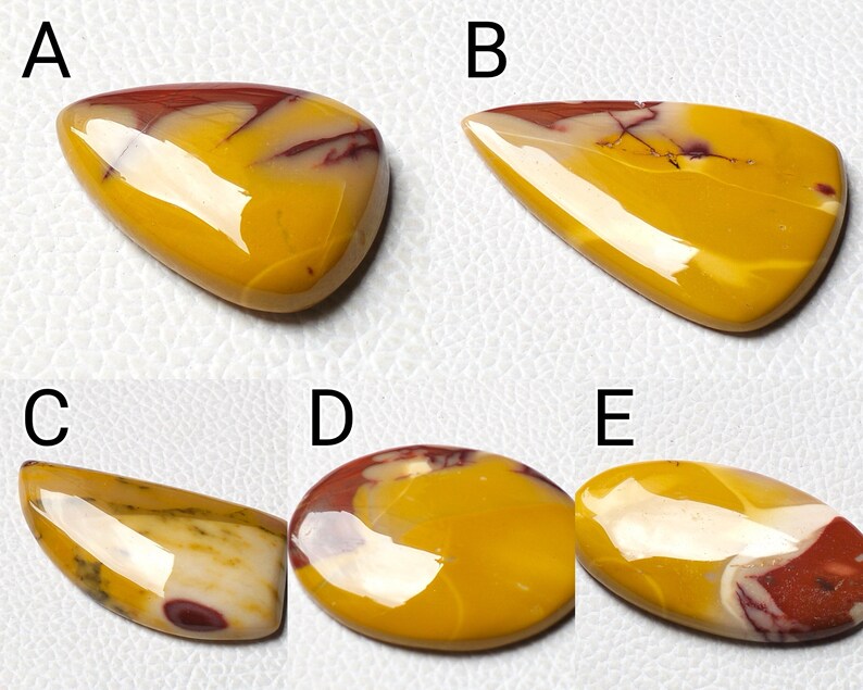 Op de afbeelding: Vijf gepolijste cabochons van gele, rode en witte edelstenen. De cabochons hebben verschillende vormen, waaronder druppelvorm, ovaal en onregelmatig.