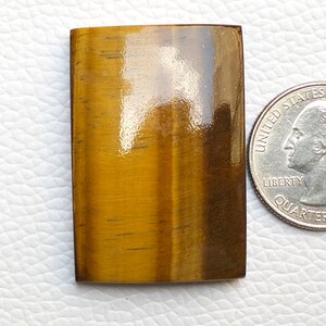 May include: A rectangular, polished tiger's eye gemstone, displaying a warm, golden-brown hue and chatoyancy. The gemstone is shown next to a U.S. quarter for size comparison.