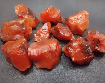 May include: A collection of ten rough, red carnelian crystals. The crystals are various sizes and shapes, with some having a darker, almost black, coloration.