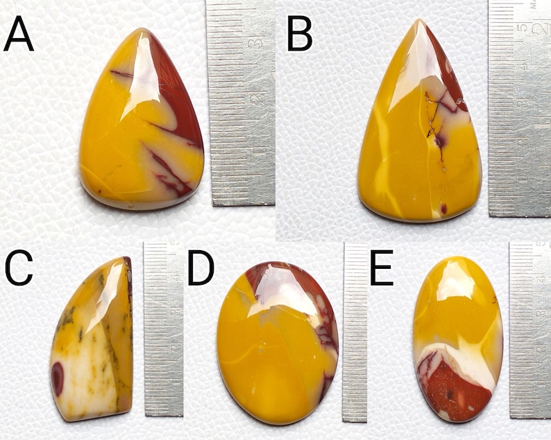 Op de afbeelding: Vijf gepolijste edelsteencabochons in verschillende vormen en maten. De stenen zijn een mix van geel, rood en wit met draaiende patronen. De stenen worden naast een liniaal getoond ter referentie van de grootte.
