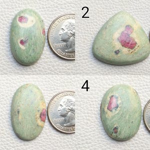 Op de afbeelding: Vier groene edelstenen met rode insluitsels worden naast een munt getoond. De edelstenen hebben verschillende vormen: ovaal, driehoekig en afgerond. De stenen hebben een lichtgroene kleur met rode en crèmekleurige insluitsels.