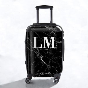 May include: Black suitcase with a white marble pattern and the initials "LM" in white. The suitcase has a retractable handle and four wheels. The text "LIVE X MAINTAIN" is printed on the bottom of the suitcase.