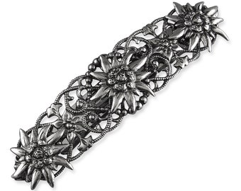 May include: A silver-toned barrette featuring a floral design with detailed edelweiss flowers. The barrette has an intricate, openwork pattern with small beaded accents and is suitable for various hairstyles.