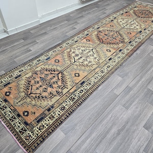 May include: A long, narrow rug with a traditional Persian design. The rug has a cream background with intricate patterns in shades of orange, brown, and black.