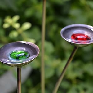 May include: Two garden ornaments with shallow, metallic bowls. One bowl contains a green glass stone, the other a red one. The stakes are set against a blurred green background, suggesting a garden setting.