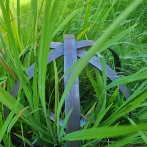 May include: A metal garden ornament, partially hidden by green grass and foliage. The ornament features a circular design with intersecting bars, creating a geometric pattern. The metal has a rustic, aged appearance.