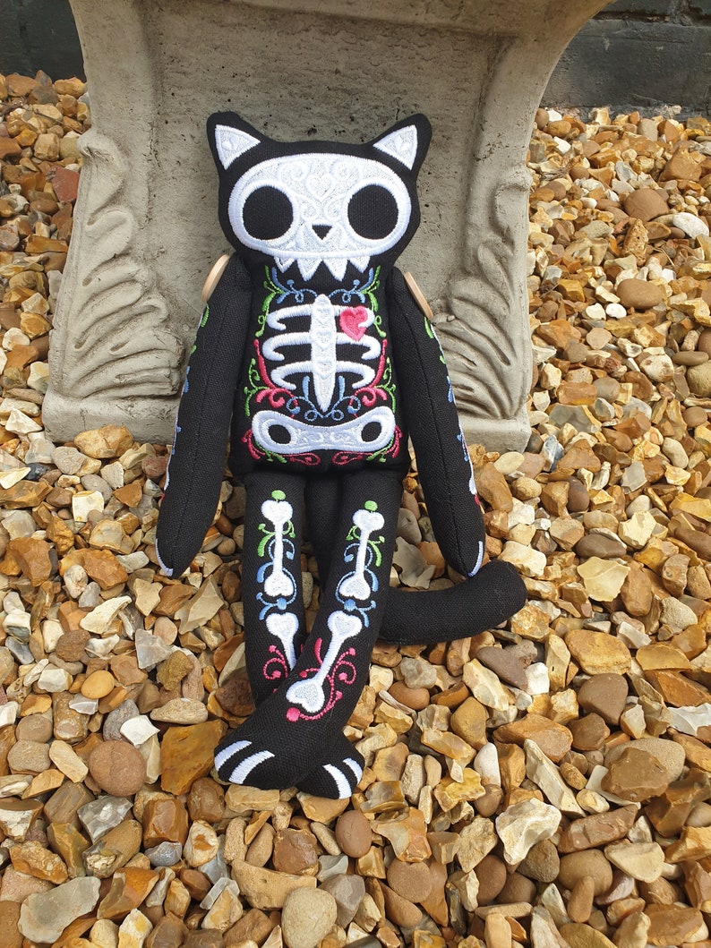 May include: A black cat skeleton plush toy with white embroidered details, including a skull, ribs, and bones. The toy has a pink heart on its chest and is sitting on a bed of small brown stones.