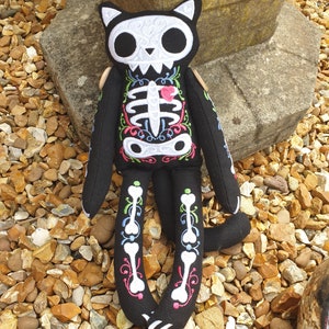 May include: Black cat plush toy with embroidered white skeleton and floral details. The cat is sitting on a bed of small stones.