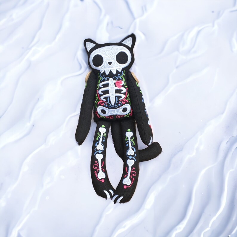 May include: A black plush cat toy with a white skeleton design and a red heart on its chest. The cat is lying on a white surface.