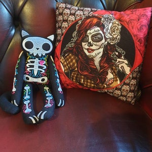 May include: A black and white skeleton cat plush toy with colorful embroidery sits on a red leather couch next to a decorative pillow featuring a woman with a sugar skull makeup design and red roses.