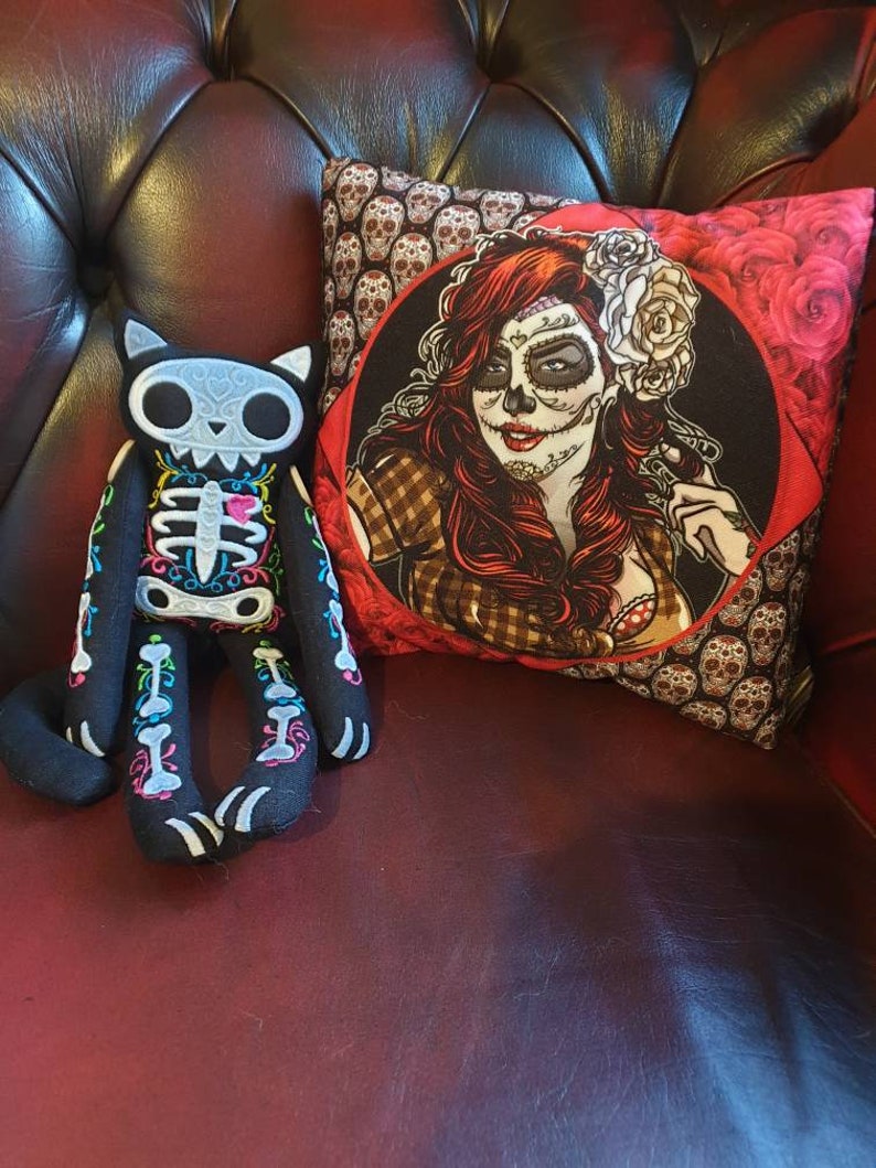 May include: A black and white skeleton cat plush toy with colorful embroidery sits on a red leather couch next to a decorative pillow featuring a woman with a sugar skull makeup design and red roses.