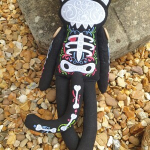 May include: Black cat plush toy with a white skeleton embroidered on its body. The skeleton is decorated with colorful floral details. The cat is lying on a bed of small brown and white stones.