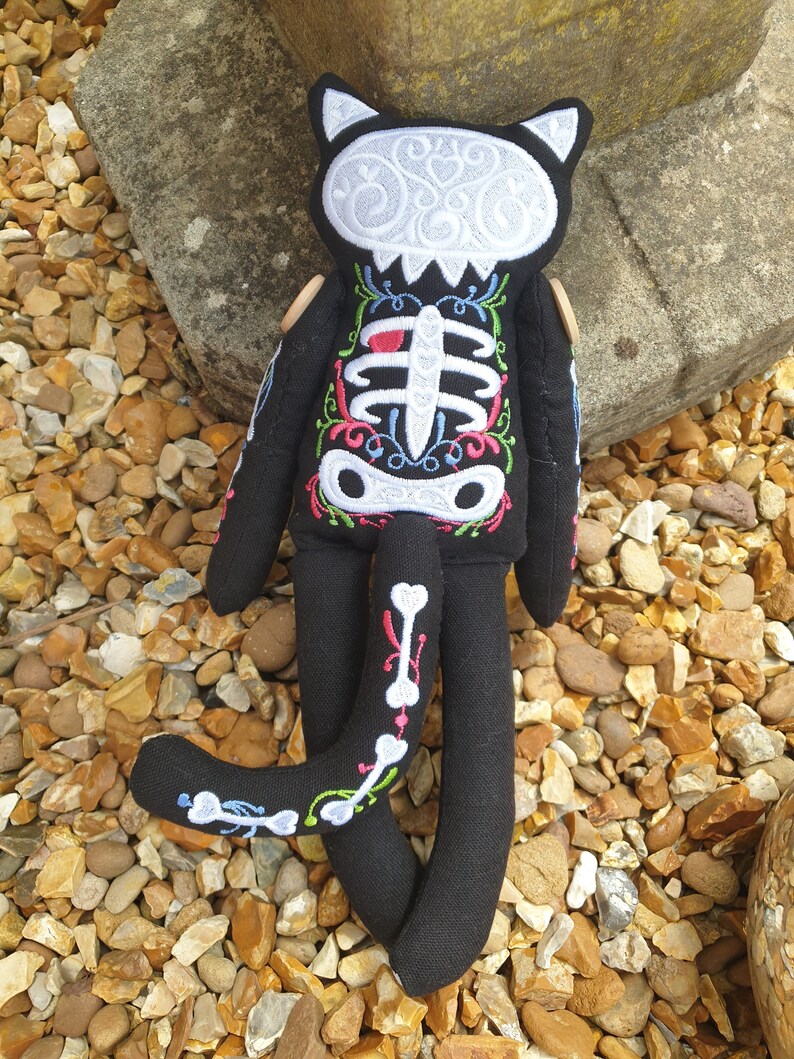 May include: Black cat plush toy with a white skeleton embroidered on its body. The skeleton is decorated with colorful floral details. The cat is lying on a bed of small brown and white stones.