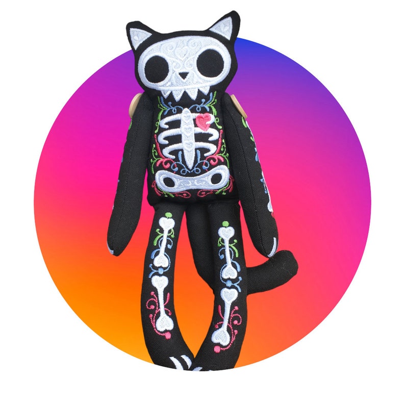 May include: A black plush cat toy with a skeleton design and colorful embroidery. The cat has a pink heart on its chest and is sitting on a gradient background.