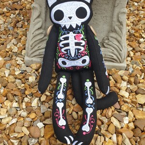 May include: Black plush cat toy with a white skeleton and floral embroidery. The cat has a pink heart on its chest and a white skull face.