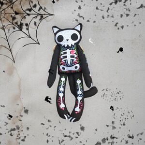 May include: Black plush cat toy with embroidered skeleton and floral details. The cat has a white skull with black eyes and a pink heart on its chest.