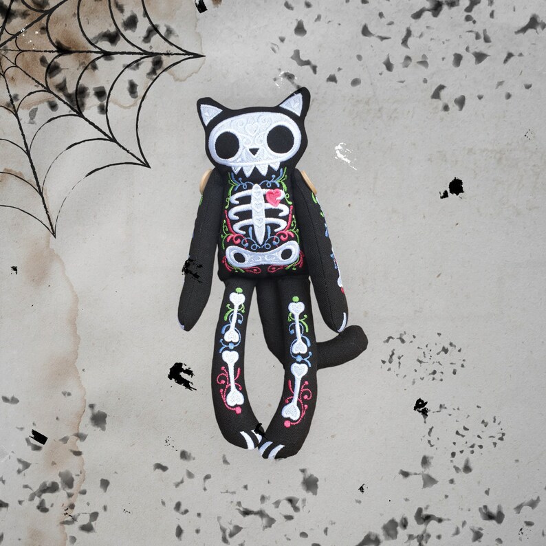 May include: Black plush cat toy with embroidered skeleton and floral details. The cat has a white skull with black eyes and a pink heart on its chest.