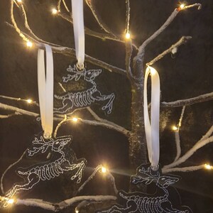 Puede incluir: Tres adornos blancos con forma de esqueleto de reno, atados con una cinta blanca, colgando de una rama nevada con luces blancas.