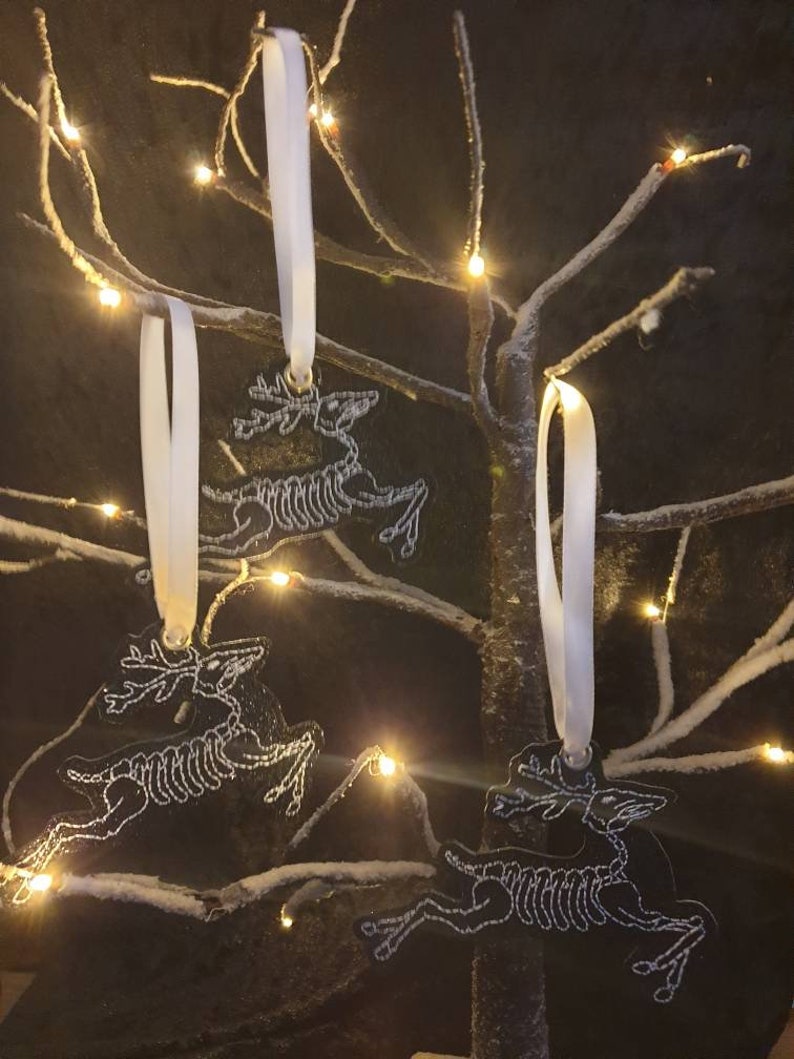 Puede incluir: Tres adornos blancos con forma de esqueleto de reno, atados con una cinta blanca, colgando de una rama nevada con luces blancas.
