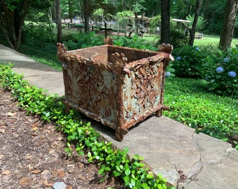 Op de afbeelding: Een grote, vierkante, roestige metalen plantenbak met ingewikkelde bloemmotieven. De plantenbak staat op een stenen pad in een tuin.
