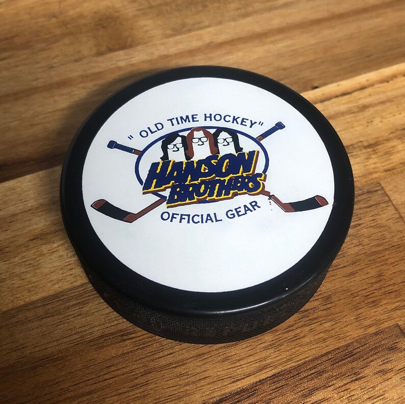 black hockey puck back side with movie logo 'hanson brothers' printed on white backing. "old time hockey" and "official gear" printed in blue.
