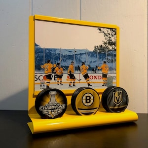 May include: Yellow hockey puck display stand with four pucks. The pucks feature logos for the Stanley Cup Champions, the Boston Bruins, and the Vegas Golden Knights. The stand has a photo of hockey players on the back.