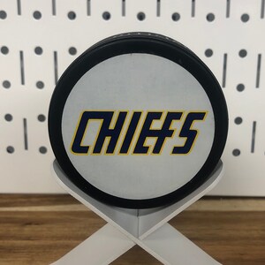 Chiefs hockey puck front side displayed on edge on a white stand that looks like crossed hockey sticks