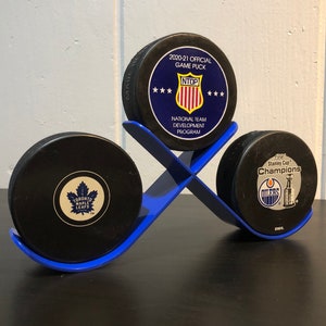 May include: Three black hockey pucks on a blue stand. The pucks feature logos for the Toronto Maple Leafs, the Edmonton Oilers, and the National Team Development Program. The Oilers puck reads "1990 Stanley Cup Champions".
