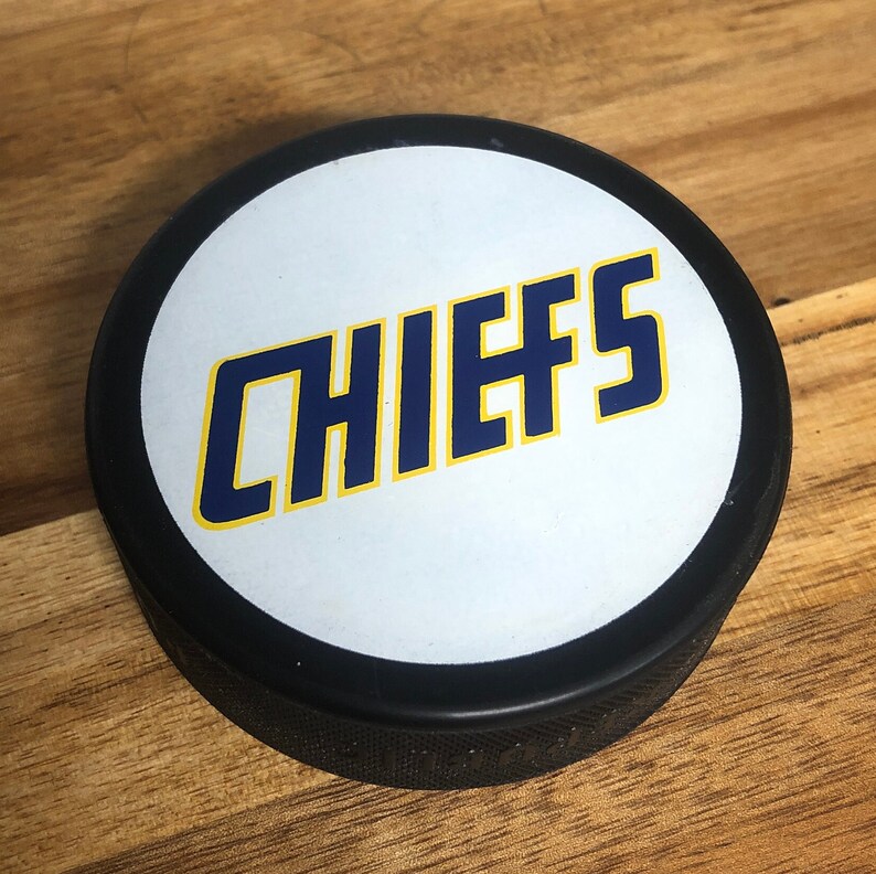 black hockey puck with movie logo 'chiefs' printed on white backing.