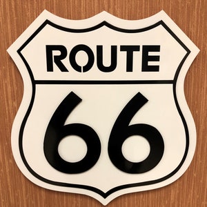 Peut inclure: Un panneau de signalisation blanc et noir Route 66 en forme de bouclier. Le panneau a une bordure noire et le texte "ROUTE 66" en lettres noires.
