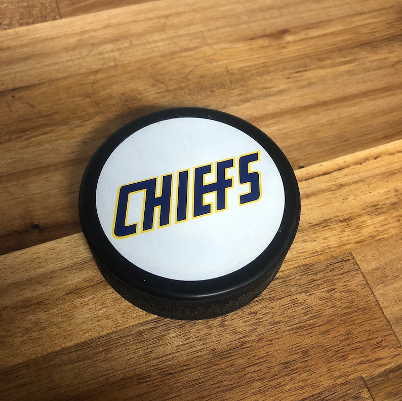 black hockey puck with movie logo 'chiefs' printed on white backing.