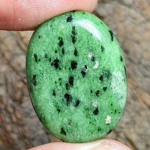Puede incluir: Una piedra preciosa de zoisita de forma ovalada con una superficie pulida y lisa. La piedra presenta un color base verde lima vibrante con inclusiones irregulares de hornblenda negra dispersas, creando un patrón moteado distintivo.