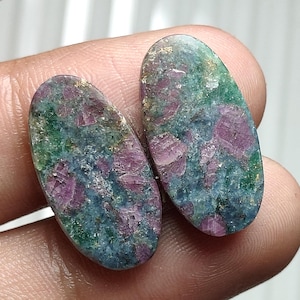 Peut inclure: Deux cabochons de forme ovale présentant un mélange tacheté d'inclusions minérales magenta profond, vert forêt et bleu ardoise. Les pierres ont une surface polie et lisse avec des textures cristallines visibles dans tout le matériau.