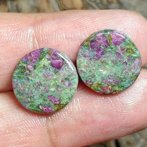 Peut inclure: Deux cabochons circulaires présentant un motif tacheté d'inclusions magenta vif et rose foncé incrustées dans une matrice vert clair et olive. Les pierres ont une surface lisse et polie avec une finition brillante et une forme ronde plate.
