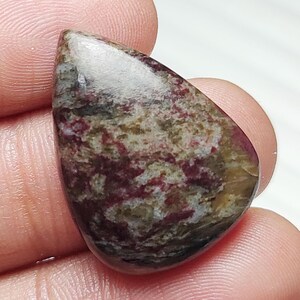 May include: A polished, teardrop-shaped gemstone with a mix of colors. The stone features a blend of burgundy, olive green, and tan hues, with a glossy, smooth surface. The gemstone is held between fingers, showcasing its natural beauty.
