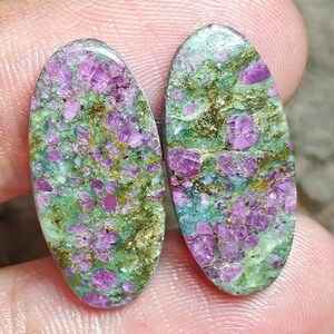 Peut inclure: Deux cabochons de forme ovale présentant un motif tacheté d'inclusions cristallines rose magenta vif incrustées dans une matrice de matériau minéral vert clair et brun terreux avec une finition de surface lisse, polie et réfléchissante.