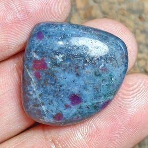May include: A polished, rounded gemstone featuring a predominantly blue and grey mottled surface with distinct, embedded inclusions of vibrant magenta and deep red spots scattered throughout the stone's smooth, reflective, and irregular shape.