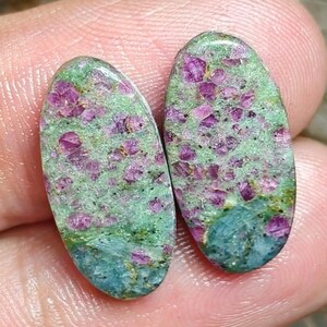 Peut inclure: Deux cabochons de forme ovale présentant un mélange d'inclusions cristallines rose magenta vif incrustées dans une matrice vert clair et bleu sarcelle. Les pierres ont une surface lisse et polie avec un contraste de couleur marqué.