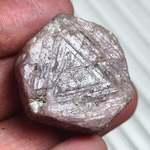 Könnte beinhalten: Ein roher, sechseckiger Edelstein in einem blassen rosa-violetten Farbton. Die Oberfläche weist ein ausgeprägtes, natürlich vorkommendes dreieckiges geometrisches Muster auf, das in die Kristallfläche eingraviert ist, mit sichtbaren Kristallstrukturen.