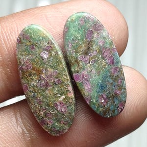 May include: Two oval-shaped gemstones with a green and brown matrix, speckled with vibrant pink and red crystals. The stones are held in a hand, showcasing their natural beauty and unique color variations.