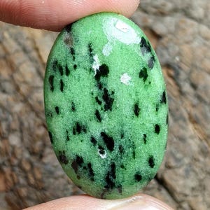 Può includere: Una gemma a cabochon di forma ovale caratterizzata da un vibrante colore di base verde chiaro con inclusioni nere irregolari sparse su tutta la superficie. La pietra presenta una finitura liscia e lucida che riflette la luce.