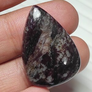 May include: A polished, teardrop-shaped gemstone with a deep burgundy base color, marbled with gray and white inclusions. The stone has a smooth, reflective surface, showcasing natural patterns and variations. The gemstone is held in a hand, highlighting its size and shape.
