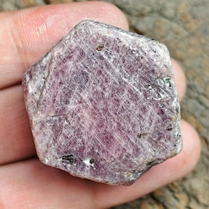 May include: A raw, hexagonal-shaped ruby crystal with a translucent, purplish-pink hue. The surface displays a natural, fibrous texture with visible striations, small dark mineral inclusions, and a slightly uneven, crystalline edge structure.