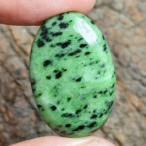 Può includere: Una pietra preziosa cabochon di forma ovale con una superficie liscia e lucida, un colore di base verde chiaro vibrante e inclusioni nere scure e irregolari sparse in tutta la pietra, creando un motivo visivo maculato ad alto contrasto.