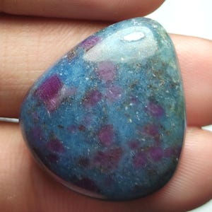 Puede incluir: Una gema pulida de forma ovalada, con una base azul-verdosa. Está salpicada de inclusiones rojo-púrpura intenso. La superficie lisa y reflectante revela patrones naturales. La gema se sostiene en una mano.