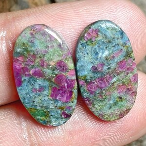 May include: Two oval-shaped cabochon gemstones featuring a mottled pattern of vibrant magenta-pink, light blue, and earthy green mineral inclusions. The stones have a smooth, polished surface that reflects light across their multicolored, textured appearance.