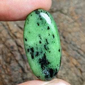 May include: An oval-shaped cabochon gemstone featuring a vibrant lime green base color with scattered, irregular black mineral inclusions throughout the surface. The stone has a smooth, polished finish that reflects light across its curved top.